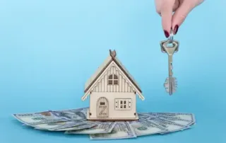 A person holding keys next to a miniture house that has money laid under it in front of the blue background representing buying a house