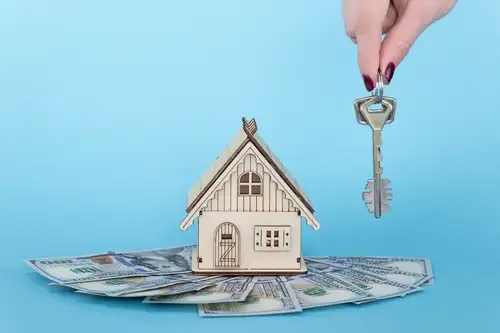 A person holding keys next to a miniture house that has money laid under it in front of the blue background representing buying a house